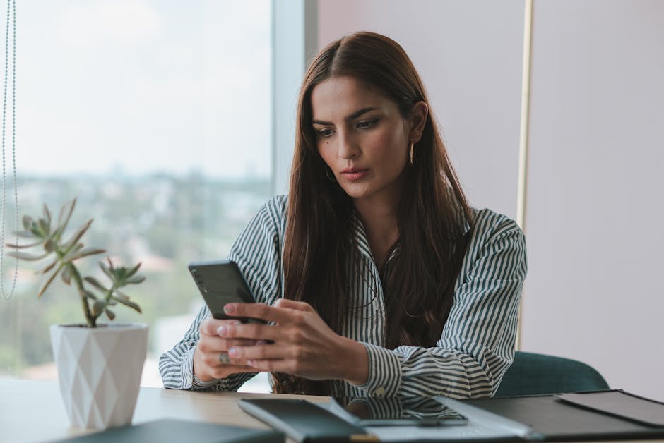 A businesswoman attentively using her smartphone at a desk in a modern office setting with a potted plant.