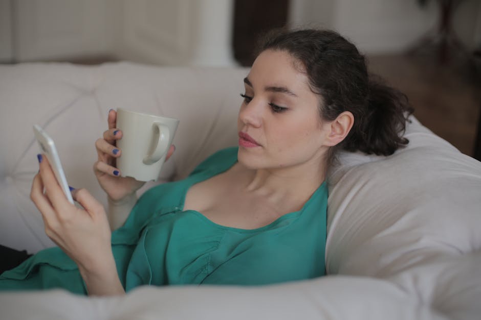 Woman sitting on a white sofa, using smartphone and holding coffee cup indoors.
