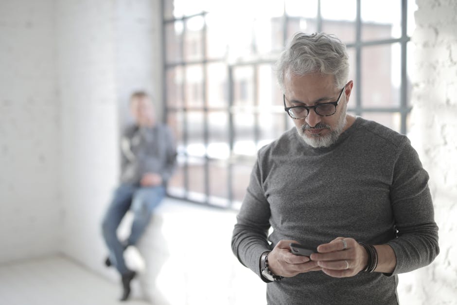 Focused mature male employee in eyeglasses and casual wear browsing internet on cellphone while standing near fenced window behind colleague in sunlight